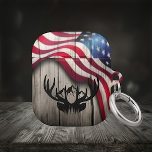 May include: A red, white, and blue American flag with a woodgrain background and a black silhouette of a deer head with antlers and mountains. The case has a silver metal ring attached.