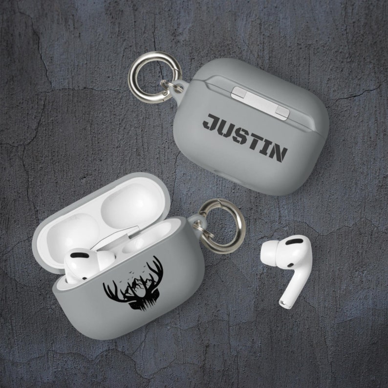 May include: Two gray Airpod cases with a carabiner clip. One case has a black deer head design with mountains and trees. The other case has the name "JUSTIN" printed on it.