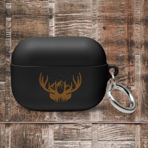 May include: Black Airpods case with a brown silhouette of a deer head with antlers and a mountain range inside the antlers. The case has a silver metal carabiner attached.