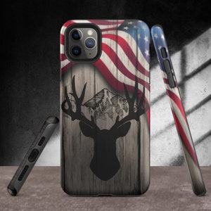 May include: A phone case with a wood grain background and a silhouette of a deer with antlers. The deer is in front of a mountain range. The top half of the case features an American flag.
