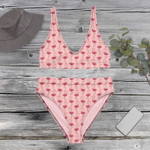 Puede incluir: Un conjunto de bikini rosa de dos piezas con un estampado de flamencos. La parte superior es de estilo cuello redondo, y la parte inferior de cintura alta presenta el mismo dise&ntilde;o de flamencos. Tambi&eacute;n se ve un sombrero de pescador gris y un smartphone.