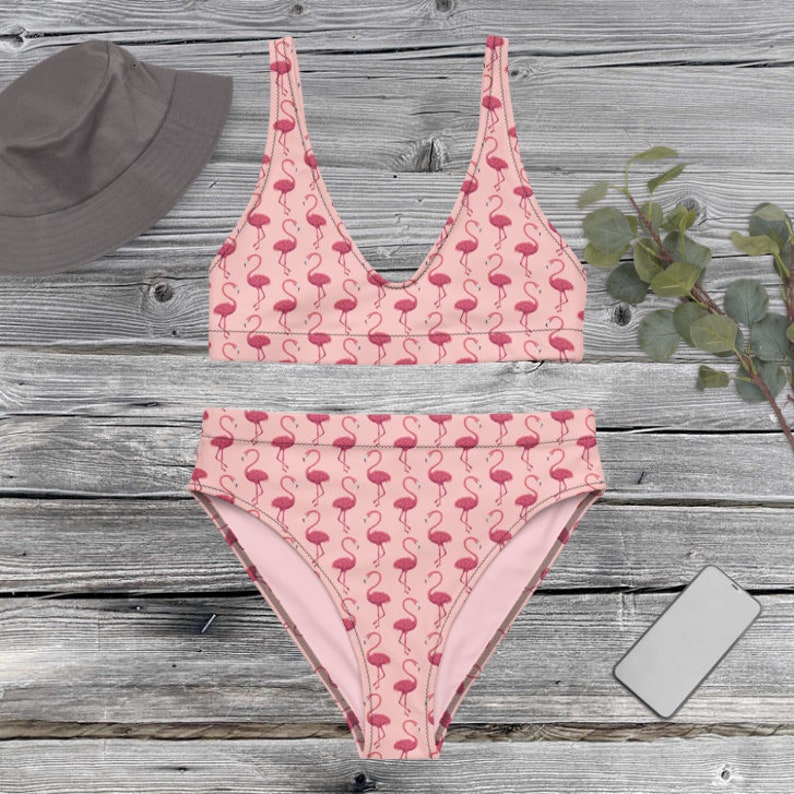 Puede incluir: Un conjunto de bikini rosa de dos piezas con un estampado de flamencos. La parte superior es de estilo cuello redondo, y la parte inferior de cintura alta presenta el mismo dise&ntilde;o de flamencos. Tambi&eacute;n se ve un sombrero de pescador gris y un smartphone.