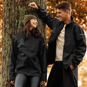 Op de afbeelding: Twee mensen dragen zwarte jassen met de tekst "Bayside" en een logo op de linkerborst. De jassen hebben een ritssluiting en zakken. Eén persoon draagt een groene muts. De jassen worden buiten gedragen in de buurt van een boom.