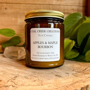 May include: A brown glass jar candle with a gold lid. The label reads "COAL CREEK CREATIONS" and "APPLES & MAPLE BOURBON". The candle is 9 oz and has a 50-hour burn time. The background includes greenery and a wooden surface.