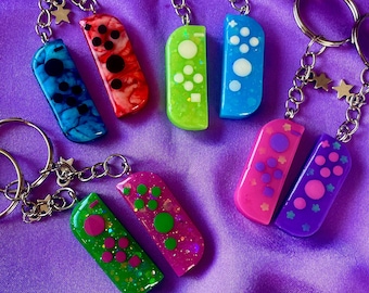 May include: A set of eight colorful keychains shaped like Nintendo Switch Joy-Cons. Each keychain features a different color and design, including blue, green, pink, purple, and red. The keychains are attached to silver key rings.