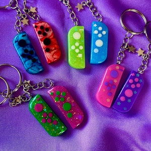 May include: A set of eight colourful keychains shaped like Nintendo Switch Joy-Cons. Each keychain features a different colour and design, including blue, green, pink, purple, and red. The keychains are attached to silver key rings.