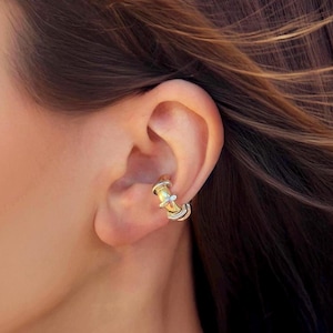 Può includere: Un ear cuff oro e argento è indossato sull'orecchio. L'ear cuff ha un design cilindrico con un centro dorato e dettagli argentati. L'ear cuff è un gioiello.