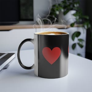 May include: A black heat-sensitive coffee mug with a large red heart design. The mug is filled with a hot beverage, and steam rises from the top. The mug's handle is black, and the interior is a light tan color.