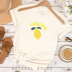 May include: A white t-shirt with a yellow lemon graphic and the word "LIMONE" in yellow letters. The shirt is on a wooden surface with sandals, a hat, and a cup of coffee.