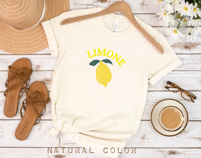 May include: A white t-shirt with a yellow lemon graphic and the word "LIMONE" in yellow letters. The shirt is on a wooden surface with sandals, a hat, and a cup of coffee.