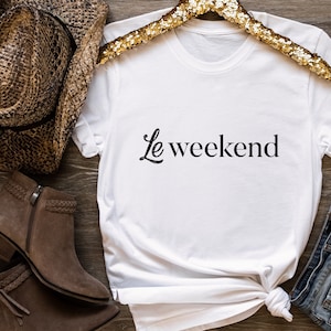 May include: White t-shirt with the text "Le weekend" in black script font. The shirt is on a wooden surface with a brown cowboy hat, brown boots, and blue jeans.