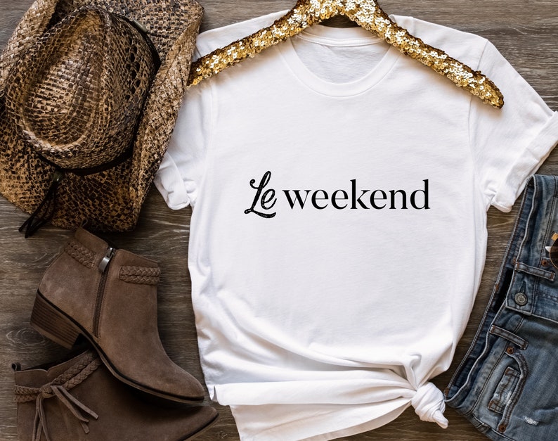 May include: White t-shirt with the text "Le weekend" in black script font. The shirt is on a wooden surface with a brown cowboy hat, brown boots, and blue jeans.