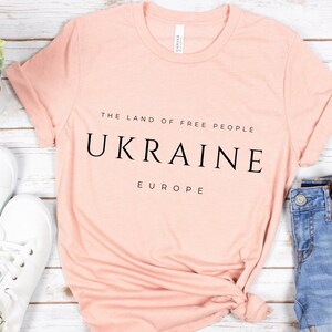 Pu&ograve; includere: Una maglietta rosa chiaro con la scritta "THE LAND OF FREE PEOPLE UKRAINE EUROPE" stampata in nero.