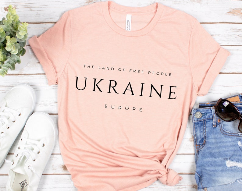 Pu&ograve; includere: Una maglietta rosa chiaro con la scritta "THE LAND OF FREE PEOPLE UKRAINE EUROPE" stampata in nero.