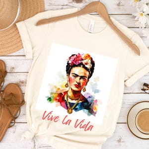 May include: Cream-colored t-shirt featuring a colorful portrait of Frida Kahlo with floral headpiece and gold jewelry. The text "Vive la Vida" is printed in red. The shirt is displayed with sandals, a hat, and a cup of coffee.