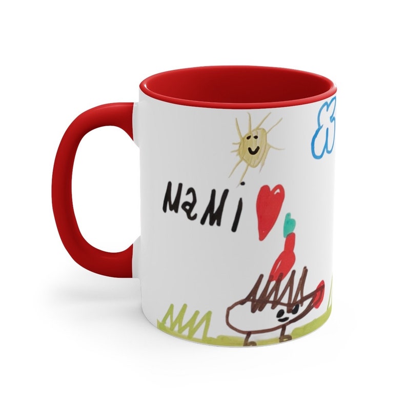 Puede incluir: Taza de cer&aacute;mica blanca con borde y asa rojos. La taza presenta un dibujo infantil de un sol, una nube, un coraz&oacute;n y un erizo. El dibujo tambi&eacute;n incluye el texto "MAMI" en marcador negro.