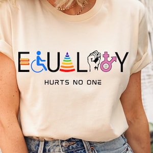 May include: A white t-shirt with the word "EQUALITY" printed in black letters. The letters are decorated with various symbols representing different groups of people, including a wheelchair, a rainbow, a fist, and a transgender symbol. The text "HURTS NO ONE" is printed below the word "EQUALITY".
