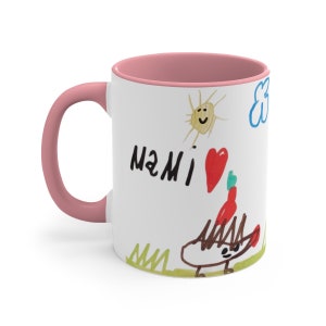 Puede incluir: Taza de cer&aacute;mica blanca con borde y asa de color rosa. La taza presenta un dibujo infantil de un sol, una nube, un coraz&oacute;n y un erizo. El dibujo tambi&eacute;n incluye las palabras "MAMI" en marcador negro.