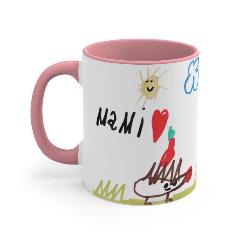 Puede incluir: Taza de cer&aacute;mica blanca con borde y asa de color rosa. La taza presenta un dibujo infantil de un sol, una nube, un coraz&oacute;n y un erizo. El dibujo tambi&eacute;n incluye las palabras "MAMI" en marcador negro.