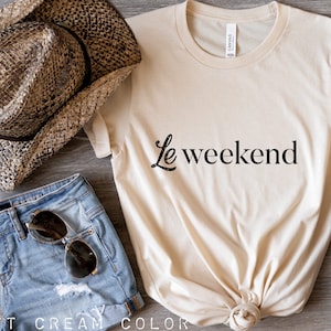 May include: A cream colored t-shirt with the text "Le weekend" in black lettering. The shirt is knotted at the bottom and is laying on a wooden surface. There is a pair of denim shorts, a straw cowboy hat, and a pair of brown suede shoes in the image.