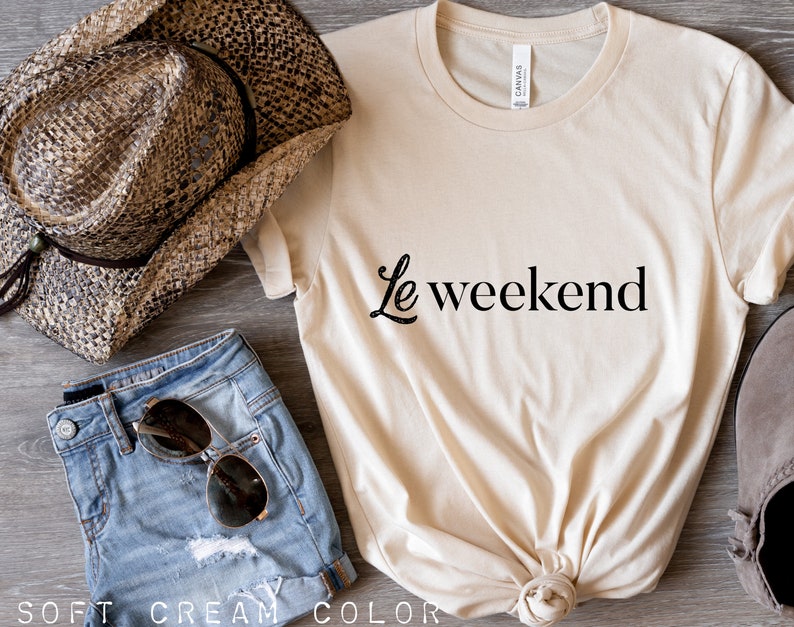 May include: A cream colored t-shirt with the text "Le weekend" in black lettering. The shirt is knotted at the bottom and is laying on a wooden surface. There is a pair of denim shorts, a straw cowboy hat, and a pair of brown suede shoes in the image.