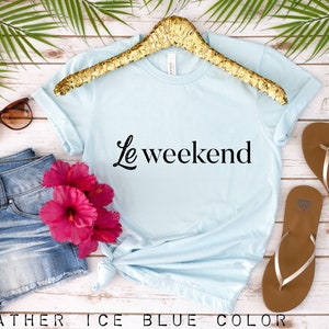 May include: A light blue t-shirt with the text "Le weekend" in black lettering. The shirt is on a hanger and there are flip flops, sunglasses, and a pair of denim shorts in the background.