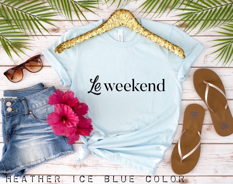 May include: A light blue t-shirt with the text "Le weekend" in black lettering. The shirt is on a hanger and there are flip flops, sunglasses, and a pair of denim shorts in the background.