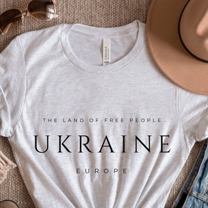 Pu&ograve; includere: Una maglietta grigia con il testo "The Land of Free People" e "UKRAINE" in grandi lettere. Il testo "EUROPE" &egrave; stampato sotto la parola "UKRAINE". La maglietta &egrave; stesa su una superficie marrone con un cappello, occhiali da sole e jeans.