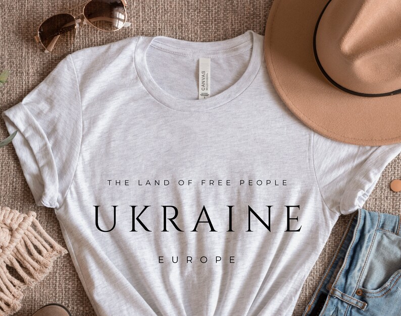 Pu&ograve; includere: Una maglietta grigia con il testo "The Land of Free People" e "UKRAINE" in grandi lettere. Il testo "EUROPE" &egrave; stampato sotto la parola "UKRAINE". La maglietta &egrave; stesa su una superficie marrone con un cappello, occhiali da sole e jeans.