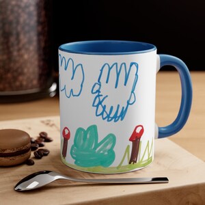 Puede incluir: Una taza de cer&aacute;mica blanca con un borde y un asa azules. La taza presenta un dibujo de un ni&ntilde;o de un arbusto verde, setas rojas y nubes azules sobre un fondo blanco.
