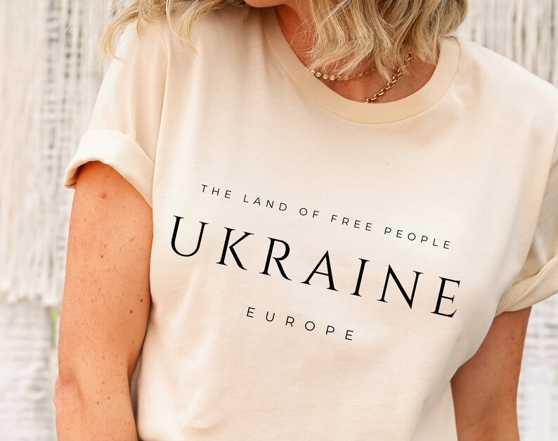 Pu&ograve; includere: Una maglietta color crema con la scritta "THE LAND OF FREE PEOPLE UKRAINE EUROPE" stampata in nero.