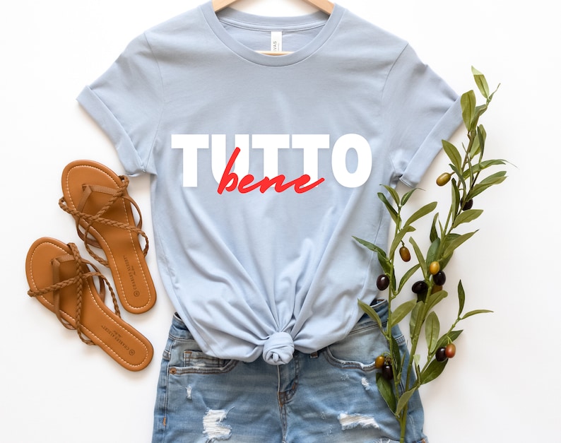May include: A light blue t-shirt with the text "TUTTO bene" in white and red lettering. The shirt is knotted at the bottom and is paired with denim shorts and brown sandals.