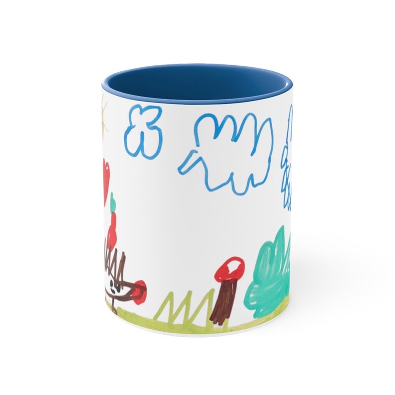 Puede incluir: Una taza de cer&aacute;mica blanca con un borde azul. La taza presenta un dibujo infantil de un &aacute;rbol verde, setas rojas, una nube azul y un animal marr&oacute;n con un coraz&oacute;n rojo.