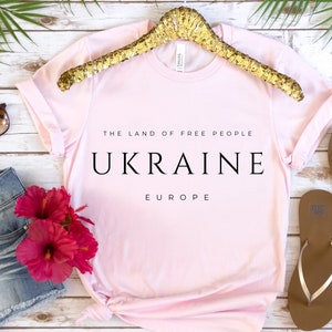 Pu&ograve; includere: Maglietta rosa con la scritta "The Land of Free People Ukraine Europe" stampata su di essa. La maglietta &egrave; su uno sfondo di legno bianco con un appendiabiti dorato, occhiali da sole, pantaloncini di jeans e infradito.