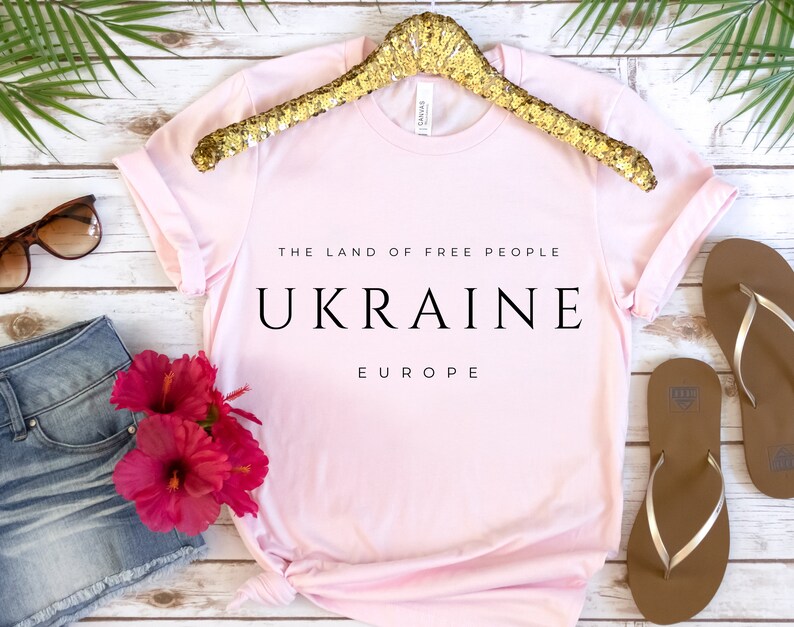 Pu&ograve; includere: Maglietta rosa con la scritta "The Land of Free People Ukraine Europe" stampata su di essa. La maglietta &egrave; su uno sfondo di legno bianco con un appendiabiti dorato, occhiali da sole, pantaloncini di jeans e infradito.