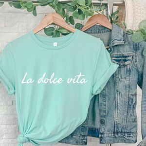 May include: A light blue t-shirt with the text "La dolce vita" in white lettering. The shirt is hanging on a wooden hanger and is paired with a denim jacket.