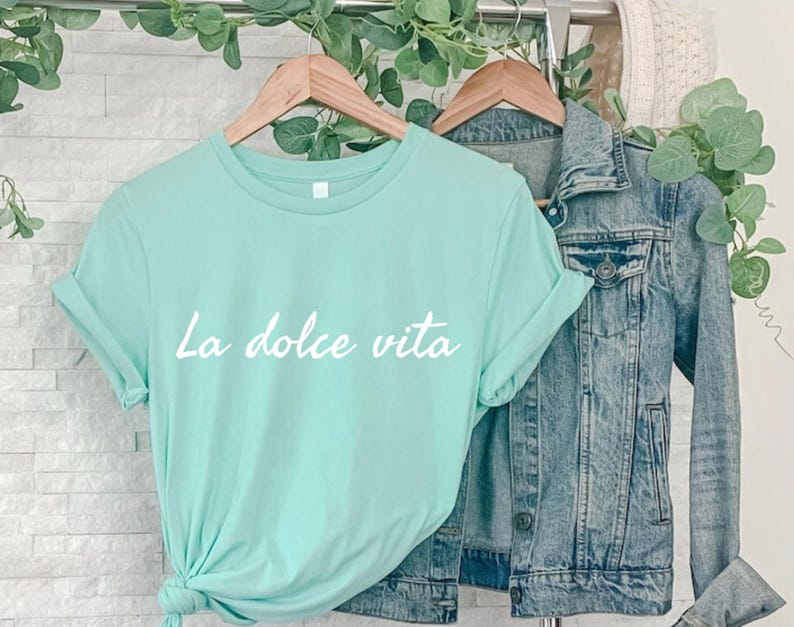 May include: A light blue t-shirt with the text "La dolce vita" in white lettering. The shirt is hanging on a wooden hanger and is paired with a denim jacket.