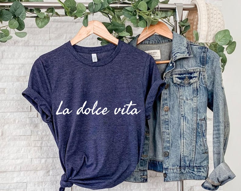 May include: A navy blue t-shirt with the text "La dolce vita" in white cursive lettering. The shirt is hanging on a hanger and is slightly creased.