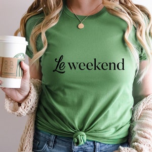 May include: A woman wearing a green t-shirt with the text "Le weekend" in black lettering. She is holding a white coffee cup with a brown paper sleeve.