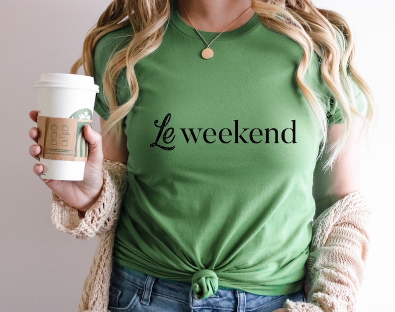 May include: A woman wearing a green t-shirt with the text "Le weekend" in black lettering. She is holding a white coffee cup with a brown paper sleeve.
