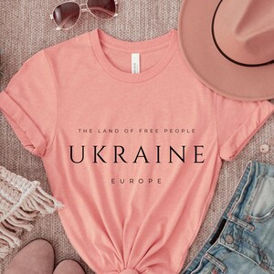 Pu&ograve; includere: Una maglietta color pesca con il testo "The Land of Free People, Ukraine, Europe" stampato in nero.