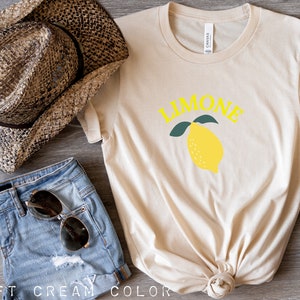 May include: A cream colored t-shirt with the word "LIMONE" in yellow letters and a yellow lemon with green leaves. The shirt is knotted at the bottom.