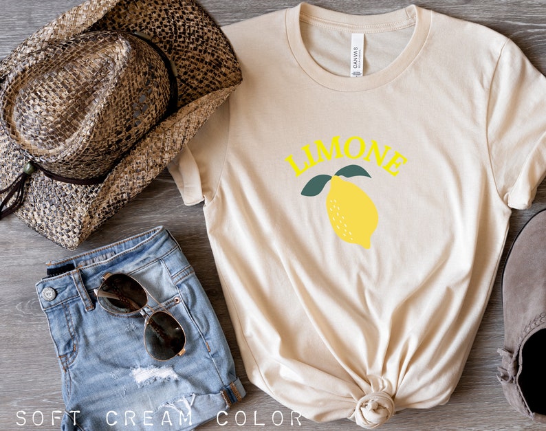 May include: A cream colored t-shirt with the word "LIMONE" in yellow letters and a yellow lemon with green leaves. The shirt is knotted at the bottom.