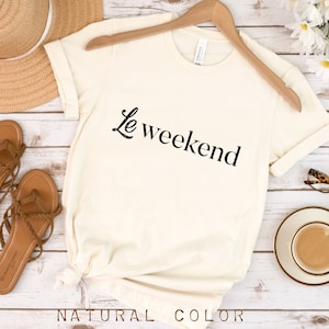 May include: A white t-shirt with the text "Le weekend" in black lettering. The shirt is on a wooden surface with a straw hat, sandals, a cup of coffee, and glasses.