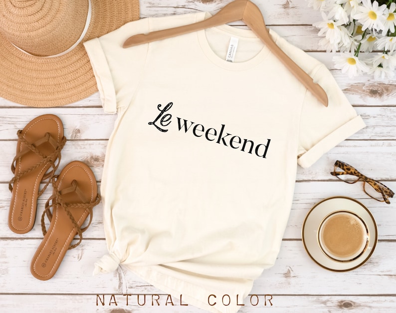 May include: A white t-shirt with the text "Le weekend" in black lettering. The shirt is on a wooden surface with a straw hat, sandals, a cup of coffee, and glasses.