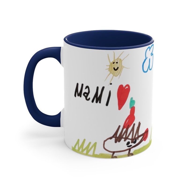 Puede incluir: Taza de caf&eacute; de cer&aacute;mica blanca con borde y asa azul marino. La taza presenta un dibujo infantil de un sol, una nube, un coraz&oacute;n y la palabra "MAMI" escrita en negro.