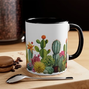 May include: White ceramic coffee mug with a black rim and handle. The mug features a colorful illustration of a variety of cacti and succulents.