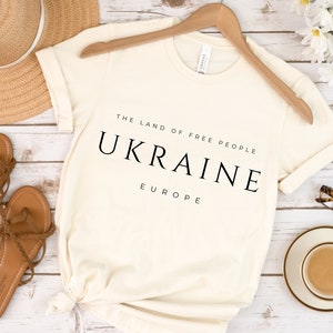 Pu&ograve; includere: Una maglietta bianca con la scritta "The Land of Free People Ukraine Europe" stampata su di essa. La maglietta &egrave; appesa a un appendiabiti di legno ed &egrave; circondata da altri oggetti, come un cappello di paglia, sandali e una tazza di caff&egrave;.