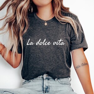 May include: A dark grey t-shirt with the text "La dolce vita" printed in white on the front. The shirt is being worn by a person with long blonde hair.