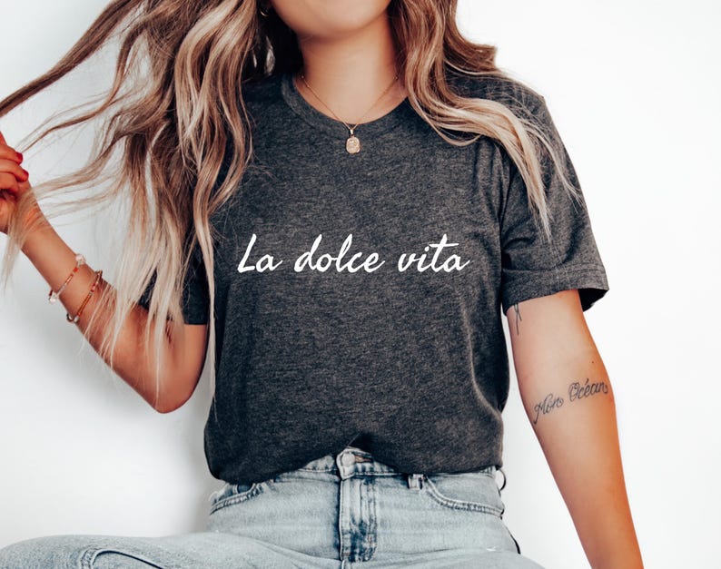 May include: A dark grey t-shirt with the text "La dolce vita" printed in white on the front. The shirt is being worn by a person with long blonde hair.